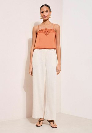 Naine kannab oranži äravõetava varrukata topi pitsdetailiga, laia säärega valgeid pükse, musti kontsaga sandaale ja kuldseid kõrvarõngaid neutraalsel taustal.