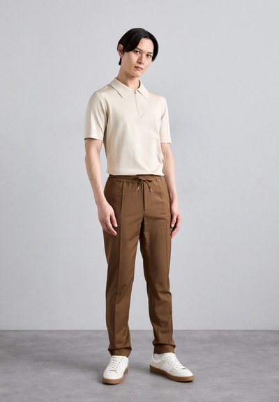 Beige zippered polo shirt, brune drawstring bukser og hvide sneakers med en tan sål. Modellen står foran en neutral baggrund.