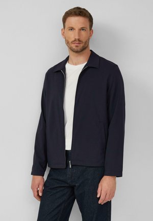 Dark navy zip-up jacket with a collared neckline and two side pockets, paired with a white t-shirt and dark denim pants. Smooth texture.
