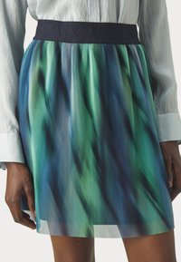 Plisserad kjol i nyanser av grönt och blått, med ett strukturerat svart midjeband. Tyget har en halvtransparent kvalitet och vertikala ränder.