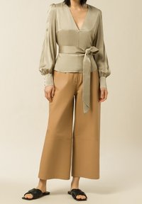 Beige Satinbluse mit Puffärmeln und einem Bindeband in der Taille, kombiniert mit weit geschnittenen, tanfarbenen Hosen und schwarzen Sandalen. Weiche Textur und elegantes Design.