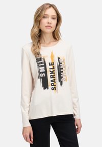 Kremowa koszulka z długim rękawem i okrągłym dekoltem, ozdobiona czarnymi, pomarańczowymi i szarymi napisami "SMILE, SPARKLE, SHINE".