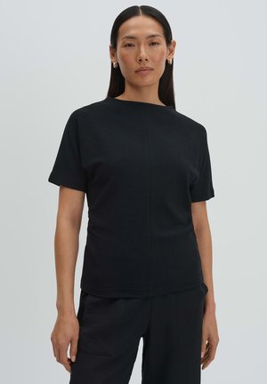 Vrouw met lang donker haar, gekleed in een zwart topje met korte mouwen en zwarte broek, staand tegen een effen lichtgrijze achtergrond.