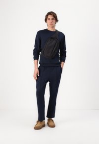 Marineblauwe sweatshirt en bijpassende broek met zichtbare naaddetails. Zwarte schoudertas en bruine schoenen met dikke rubbere zolen. Eenvoudig, modern ontwerp.