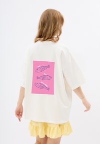 Weißes übergroßes T-Shirt mit einem pinken Grafikdruck auf dem Rücken, der drei illustrierte Fische und den Text "seas the Moment" zeigt. Kurze Ärmel.