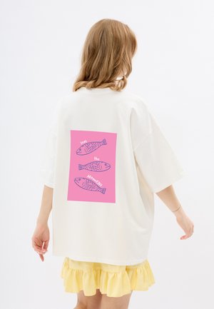 Witte oversized t-shirt met een roze graphic op de achterkant, waarop drie geïllustreerde vissen staan en de tekst "seas the Moment." Korte mouwen.