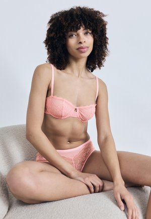 Młoda kobieta z kręconymi włosami siedząca ze skrzyżowanymi nogami, ubrana w brzoskwiniowy koronkowy biustonosz i dopasowane majtki, na jasnoszarym fotelu na tle białej ściany.