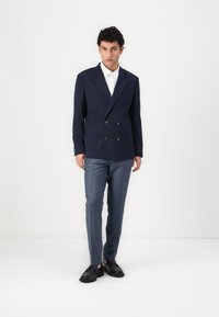 Marineblaues, doppelt gefüttertes Sakko mit zwei vorderen Taschen, kombiniert mit hellblauen, strukturierten Hosen und schwarzen, klobigen Schuhe.