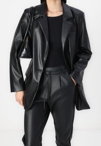 Vrouw in een zwarte neplederen blazer en broek, zwarte top, met een zwart schoudertas met een gouden rits.