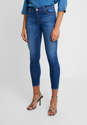 Jeans in denim blu skinny con vita alta, leggere sfumature, cinque tasche e cuciture abbinate. Abbinati a tacchi neri con cinturini.