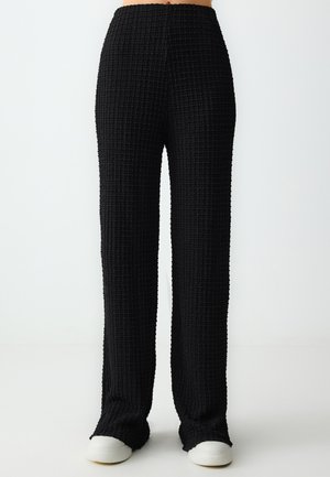 Schwarze, strukturierte, weit geschnittene Hosen mit hoher Taille und elastischem Bund. Der Stoff weist ein gitterartiges Muster auf. Kombiniert mit weißen Sneakers.