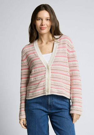 Frau trägt eine rosa-weiß gestreifte Strickjacke über einem weißen Oberteil und blauen Jeans, steht vor einem einfarbigen grauen Hintergrund.