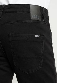 Black denim jeans feature a rear pocket with a visible stitched design and a leather patch labeled "CARS" at the waistband.