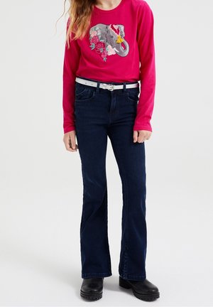 Fuchsia långärmad skjorta med elefant- och blommöster, kombinerad med mörkblå utsvängda jeans och ett vitt bälte. Svarta ankelstövlar kompletterar outfiten.