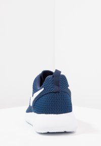 Tummansininen verkkomateriaali urheilukengät valkoisella pohjalla nähtynä takaa, jossa on vetotuki ja valkoinen logo sivulla.