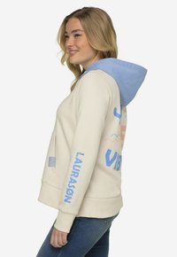 Cremefarvet hoodie med en lyseblå hætte, prydet med blå og pink "VIBRATIONS" tekst. Lavet af blødt stof med en afslappet pasform og sidelomme.