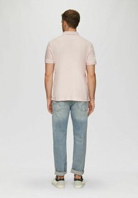 Mann steht mit dem Rücken zur Kamera, trägt ein hellrosa Polo-Shirt, hochgekrempelte, ausgebleichte Jeans und weiße Sneakers mit dunklen Akzenten.