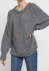 Maglione grigio a maglia con scollatura ampia, vestibilità oversize e polsini a coste. Texture morbida con motivi di cucitura visibili.