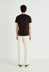 Brown short-sleeve t-shirt, white cargo pants with back pockets, brown shoes. Model faces away against a plain light background.