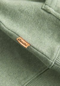 Sudadera de forro polar en verde apagado. Presenta puños y dobladillo acanalados. Incluye una etiqueta rectangular naranja con texto negro. Textura suave.