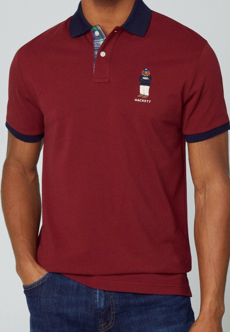 Burgundy paita polo kanssa tummansinisellä kauluksella ja ruudullisella vuorilla, jossa on brodeerattu karhulogo ja "HACKETT"-teksti rinnassa.