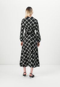 Femme aux longs cheveux blonds, portant une robe midi à manches longues noire et blanche à motifs, nouée à la taille, se tenant face à un mur blanc.
