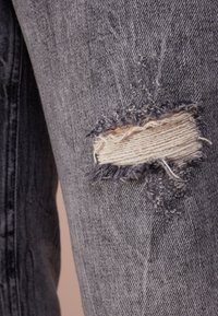 Jeans de mezclilla gris con detalles desgastados y un parche roto que expone la tela blanca de abajo. La textura es áspera con bordes deshilachados.