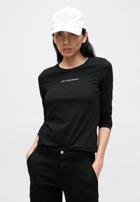 Svart langermet topp med rund hals og hvitt logotrykk. Kombinert med svarte bukser, aksessert med en hvit caps med logo.