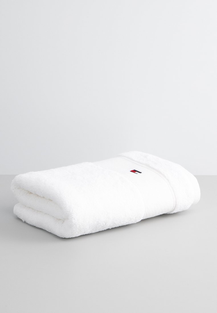 Witte katoenen handdoek, netjes opgerold, met een gestructureerd oppervlak. Het heeft een horizontale band met een klein logo in rood en blauw.