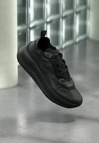 Μαύρο αθλητικό παπούτσι Calvin Klein με παχιά σόλα και κορδόνια, αιωρείται πάνω από ένα γυαλισμένο δάπεδο από μπετό κοντά σε έναν τοίχο από γυάλινους κύβους.