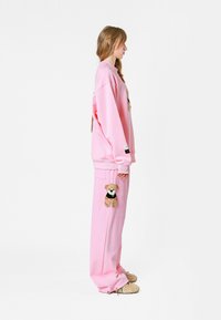 Sudadera de algodón rosa con un parche de oso de peluche en la parte delantera, combinada con pantalones rosa a juego que tienen un parche de oso de peluche en el muslo.
