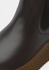 Bruine leren enkelboot met elastische zijpanelen, gestructureerde rubberen zool, ronde neus en gestikte details langs de bovenrand.