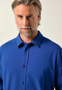 Modrá polo košile se strukturovanou tkaninou, klasickým límečkem a léga s fialovými knoflíky. Zaměření na úzký střih a jednobarevný design.
