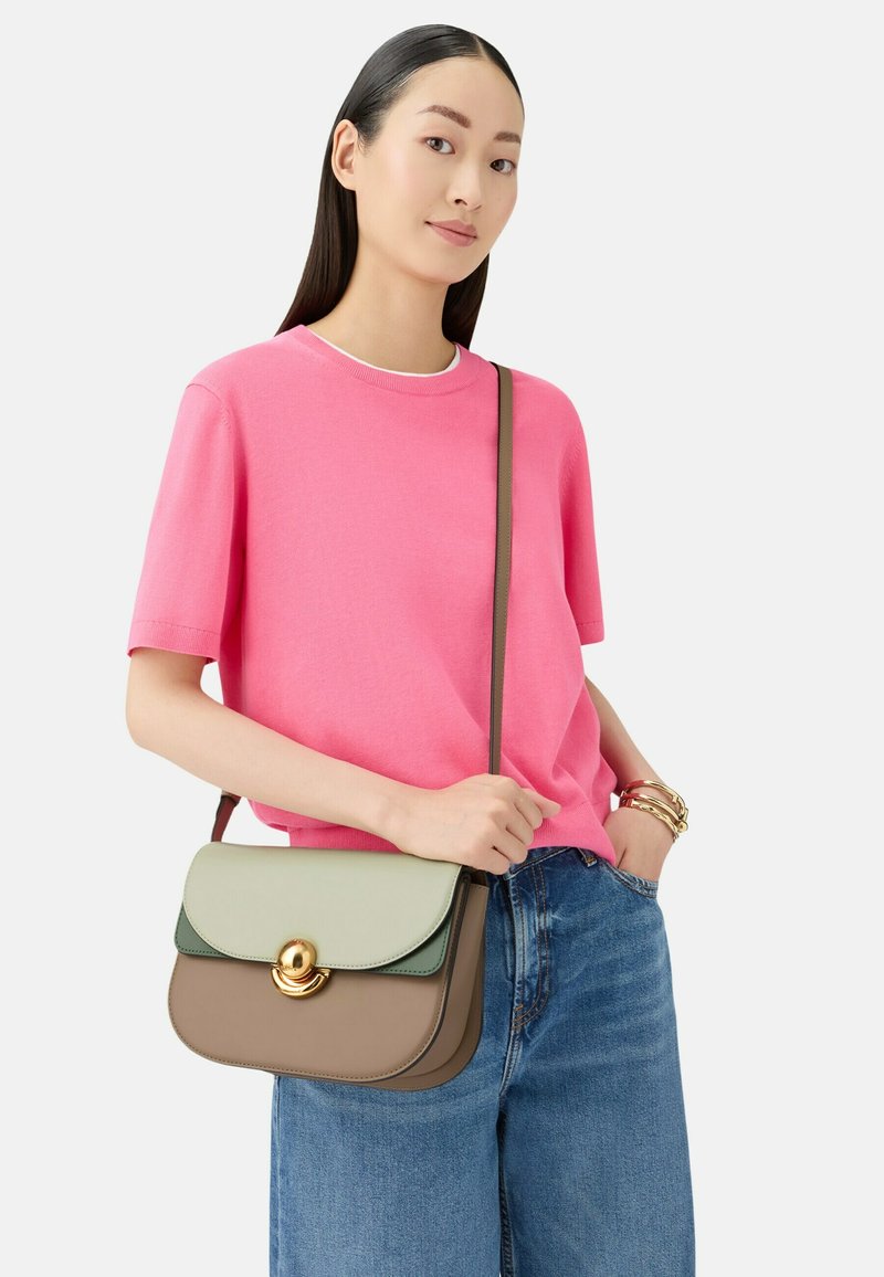 Torebka typu crossbody o zielono-brązowym skórzanym wzorze z złotym okrągłym zapięciem. Stylizowana z niebieskimi dżinsami i różową koszulą.