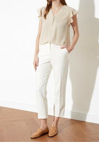 Beige Bluse mit Rüschensleeves und V-Ausschnitt; weiße, taillierte Hose mit Taschen; hellbraune Loafers mit Kettenakzent.