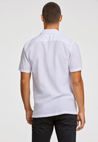 Camisa de lino blanca de manga corta con una textura suave, un cuello clásico y un dobladillo curvado, combinada con pantalones oscuros. La vista trasera muestra los detalles del diseño.