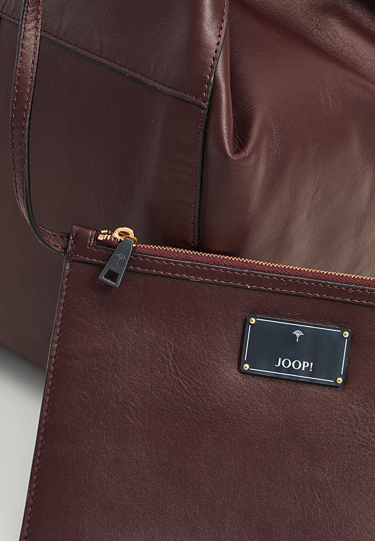 Burgunderfarbene Lederhandtasche mit glatter Textur, ausgestattet mit einer abnehmbaren Tasche, goldenen Beschlägen und einem markierten Schild mit dem eingravierten "JOOP!".
