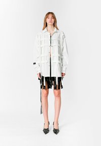 Camisa blanca de gran tamaño con un patrón de cuadrícula y acentos de cinta negra; combinada con una falda negra con tiras colgantes; tacones de punta negros.