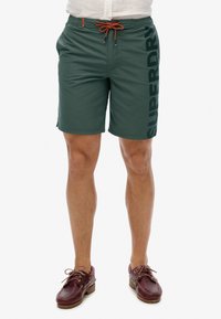 Grüne Schwimmshorts mit einem orangefarbenen Kordelzug, ausgestattet mit einem seitlichen Logo in Schwarz. Hergestellt aus leichtem Material, mit glatter Textur, bis zur Mitte des Oberschenkels.