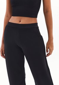 Černé sportovní kalhoty s hladkou texturou, elastickým pasem, bočními kapsami a jemným bočním pruhovým detailem.