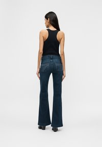 Γυναίκα με μακριά σκούρα μαλλιά που φοράει μαύρο αμάνικο τοπ, σκούρα τζιν με καμπάνα και ραμμένες τσέπες στο πίσω μέρος, καθώς και μαύρα παπούτσια με τακούνι, στέκεται γυρισμένη προς τα πίσω.