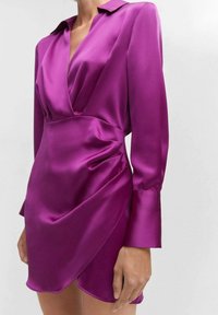 Frau, die ein figurbetontes, langärmeliges Satin-Kleid mit tiefem V-Ausschnitt in lebhaftem Lila trägt, mit einem Wickelrock und strukturierten Schultern.