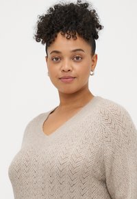 Beige V-halsgenser med et teksturert sikksakkmønster, korte ermer og et mykt, lett strikkemateriale. Subtil glans gjennom hele stoffet.