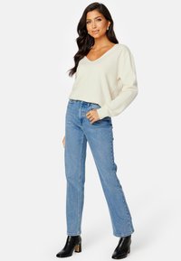 Cremiger V-Ausschnitt Oversized-Pullover, kombiniert mit hochgeschnittenen hellblauen Jeans und schwarzen, hochhackigen Stiefeletten. Weiche Textur, lässige Passform.