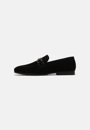 Black suede loafers with a low heel, featuring a fabric strap and decorative silver buckle. Smooth texture, pointed toe design.