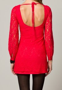 Femme portant une robe en dentelle rouge ajustée à manches longues, un dos ouvert avec un lien au cou, et des collants noirs.