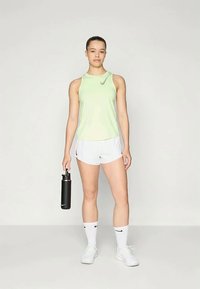 Noor naine helesinises ilma varrukateta topis ja valgetes lühikestes pükstes hoiab musta Nike veepudelit, seistes ühetoonilise tausta ees.