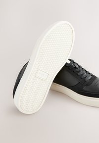 Baskets noires avec une tige en cuir lisse, une semelle en caoutchouc texturée, des accents crème et des lacets plats. Présentent des perforations sur l'avant-pied pour la respirabilité.