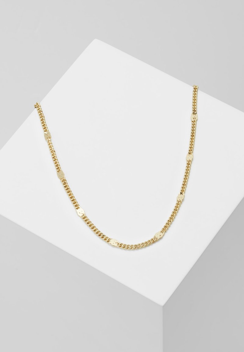 Collier chaîne en or avec des maillons décoratifs plats et ronds exposé sur un piédestal géométrique blanc sur fond gris.
