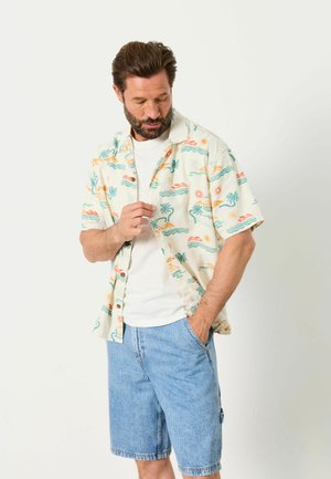Man with beard wearing a white tropical print short-sleeve shirt over a white t-shirt and blue denim shorts, hand in pocket, looking down.